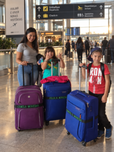 Home 48 Foto de família durante uma viagem, com malas de viagem personalizadas em destaque. Cada mala possui uma faixa elástica de mala com nome bordado, funcionando como identificador de bagagem no aeroporto. A cena transmite união familiar, organização, praticidade e estilo para viajar.